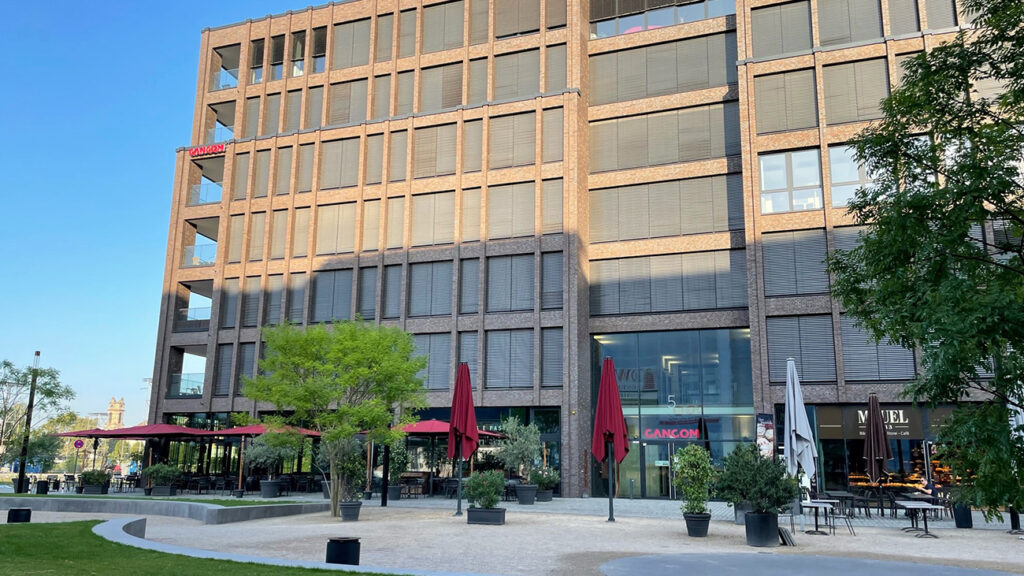 Auf dem Bild ist das moderne Büro-Gebäude von CANCOM in Köln-Mühlheim zu sehen. Im Erdgeschoss befinden sich verschiedene Gastronomie-Möglichkeiten mit Sitzplätzen und Sonnenschirmen. Davor erstreckt sich ein großer, weitläufiger Platz mit einigen Bäumen.