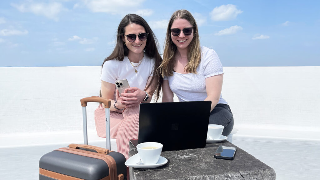 Zwei junge Frauen sind im Urlaub, sitzen mit Laptop und Smartphone in einem Café und genießen die Sonne. Ein Koffer steht neben dem Tisch.
