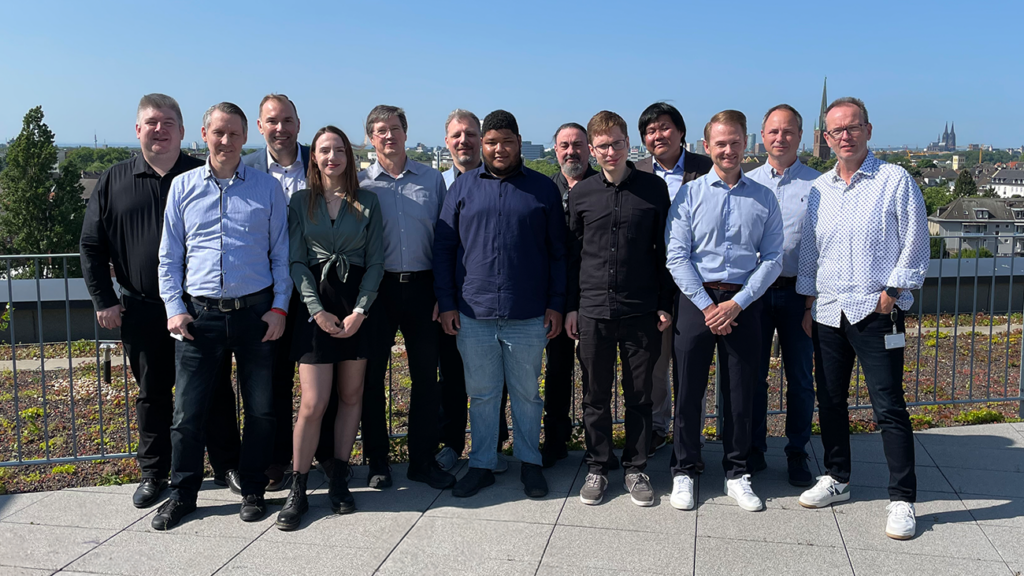 Auf dem Bild ist das komplette DPI-Team zu sehen. Sie stehen auf der Dachterrasse vom Kölner CANCOM Büro. Im Hintergrund ist der Kölner Dom zu sehen.