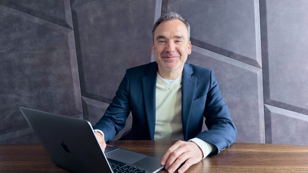 Sven sitzt an einem Tisch mit Laptop. Er trägt ein dunkelblaues Sacko und hat kurze, graue Haare.