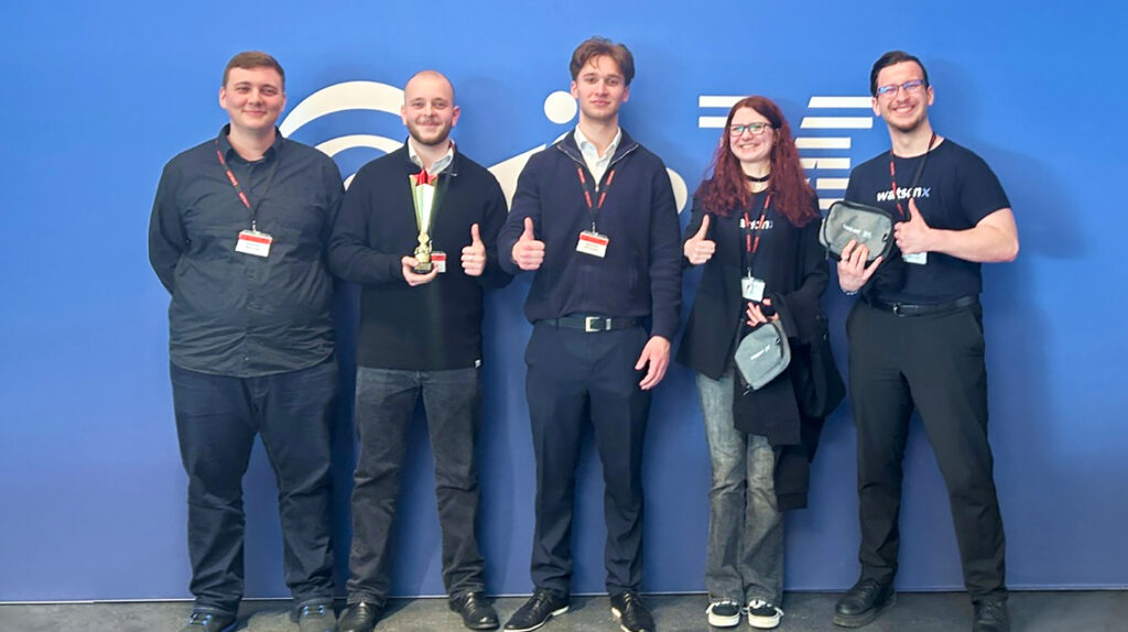 Auf dem Bild ist das Team, das bei der watsonX Challenge mitgemacht und gewonnen hat, zu sehen. Es steht vor einer blauen IBM Wand .