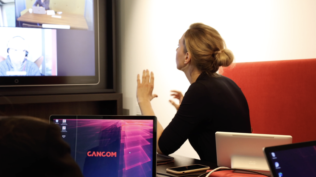 Das Bild zeigt unsere Gastautorin Catharine Hack sitzend von hinten. Sie ist in einem Online-Meeting und schaut auf einen Bildschirm, auf dem andere Teilnehmer des Meetings zu erahnen sind. Der Raum ist modern eingerichtet mit einem roten Sofa. Auf dem Tisch stehen Tablet und Laptop.