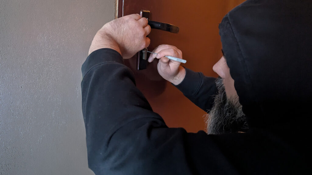 Philipp kniet vor einer Tür und versucht sie aufzubrechen. Dabei trägt er einen schwarzen Hoodie.