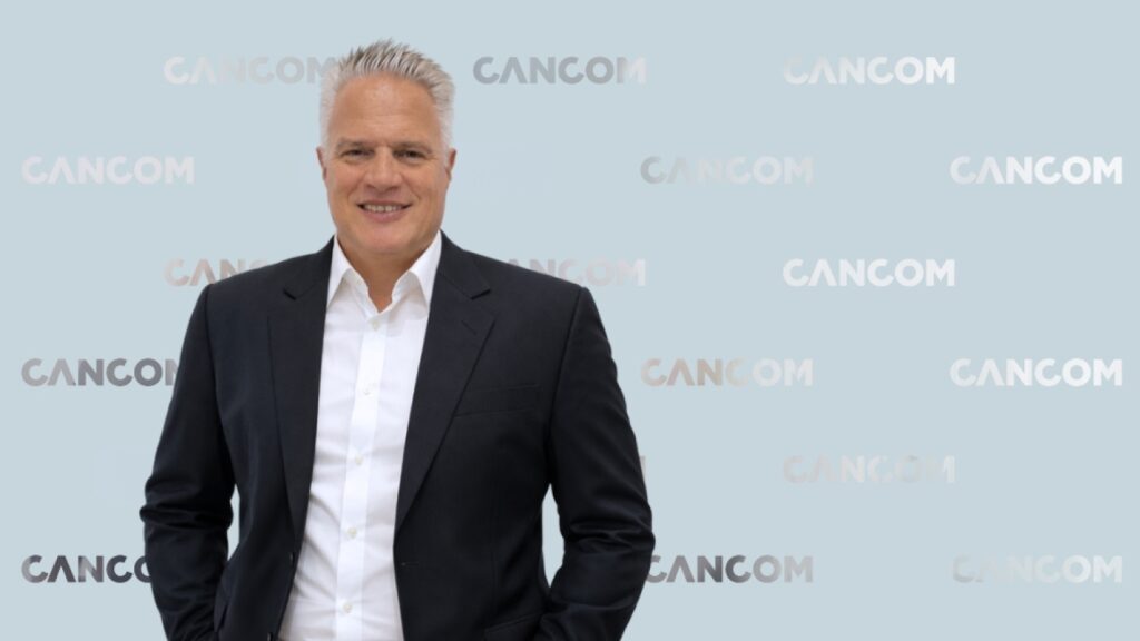 Andreas steht vor einer CANCOM gebrandeten Glaswand. Er hat graue Haare, trägt eine weißes Hemd und Sacko.