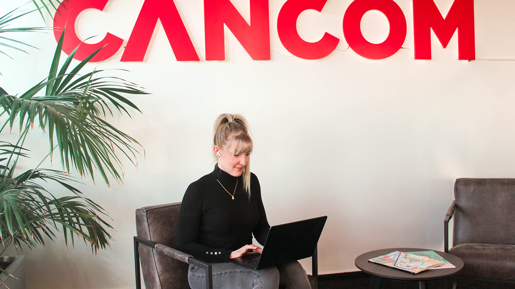 Eva sitzt auf einem Sessel, den Laptop auf den Schoß. Sie hat Kopfhörer im Ohr und schaut auf ihren Bildschirm. An der Wand hinter ihr ist das CANCOM Logo in großen, roten Buchstaben zu sehen.