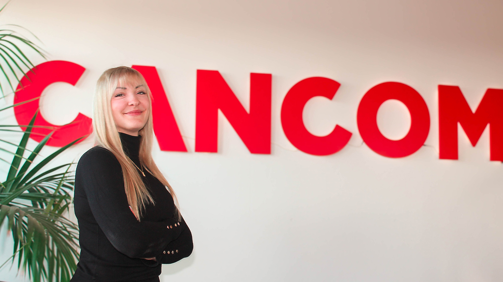 Eva Dölle steht im Büro vor einem großen, roten CANCOM Logo. Sie hat lange, blonde Haare, trägt einen schwarzen Pullover und hat die Arme vor der Brust verschränkt.